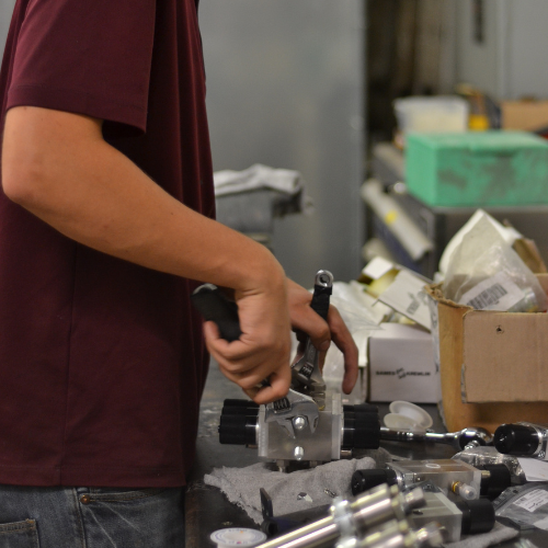 A person in a maroon t-shirt and jeans working at a cluttered workbench, wrenching on some Sames Parts. 
